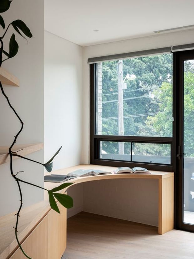 Bright, compact home office nook with a built-in timber desk beneath a large black-framed window and adjacent glazed door. Open books rest on the desk, while integrated pale timber shelving and cabinetry frame the space. A climbing indoor plant adds a natural element, and the room has light walls, timber flooring, and a clean contemporary feel.
