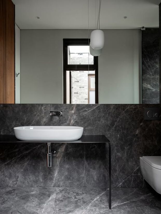 Contemporary bathroom featuring dark veined stone wall and floor surfaces, a floating black vanity with a white vessel basin, wall-mounted chrome tapware, a wall-hung toilet, black-framed window, and a suspended white pendant light. Timber cabinetry is visible at the left edge.