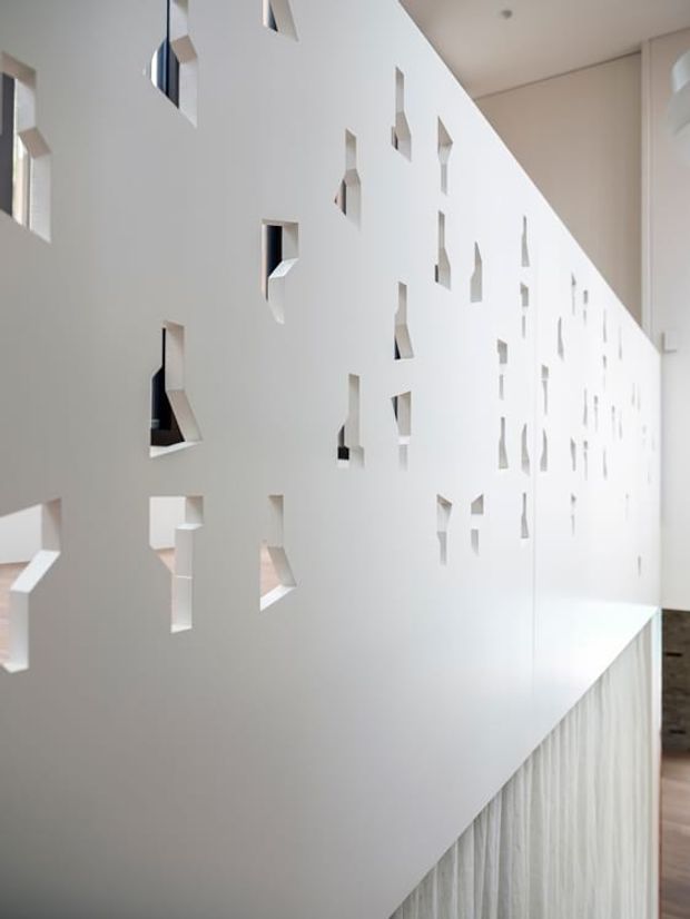 A bright, minimalist interior featuring a long white perforated partition wall with irregular geometric cut-outs. The wall overlooks a stair or lower-level void with a vertically textured white balustrade or screen, creating a sculptural architectural feature. A small glimpse of adjacent timber flooring and dark stone or tiled wall is visible at the far right.