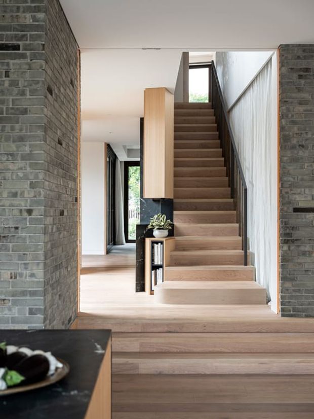 Interior view focused on a contemporary stairway rising through an open doorway. The staircase and surrounding floors are finished in warm timber, with a slim dark metal balustrade and integrated timber-and-dark cabinetry beneath the stairs. Exposed grey brick walls frame the opening, while white walls and a narrow skylit upper landing create a bright, refined transition space. A partial dark stone kitchen or bench surface is visible in the foreground.