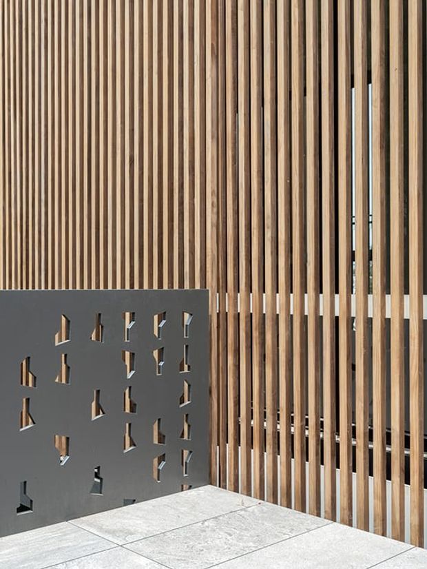 Architectural detail showing a tall vertical timber batten screen with varied natural wood tones, paired with a dark grey perforated privacy or balustrade panel. A pale large-format tiled surface occupies the foreground, and glimpses of glazing or interior space are visible through the timber slats. The composition is contemporary and materially focused.