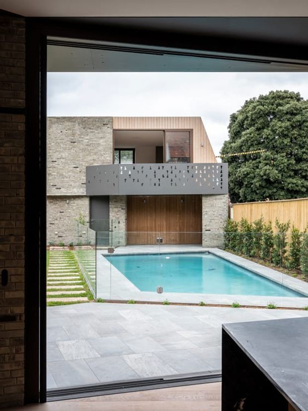 View from inside through a large black-framed opening toward a contemporary backyard courtyard. The scene features a rectangular swimming pool, pale stone paving, stepped lawn, timber fencing, a mature tree, and a modern two-storey brick-and-timber house with a perforated grey balcony and large timber entry door. A dark countertop or island edge is visible in the foreground.