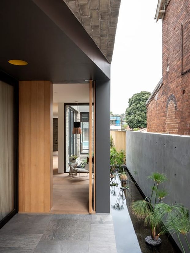 A contemporary residential entry passage with a dark soffit and charcoal structural framing, warm timber wall panelling and a glazed doorway leading into a light-filled interior. Large-format grey floor tiles continue beside a narrow reflective water feature planted with grasses. An adjoining exposed red-brick building and concrete boundary wall create a layered urban architectural setting.