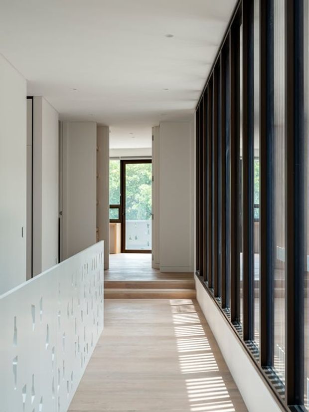 A bright, minimalist interior hallway or landing with pale timber flooring, white walls and built-in cabinetry. A perforated white balustrade borders the stair opening on the left, while a full-height wall of dark-framed glazing on the right casts strong linear sunlight across the floor. The corridor steps up toward a glazed doorway with greenery visible beyond.
