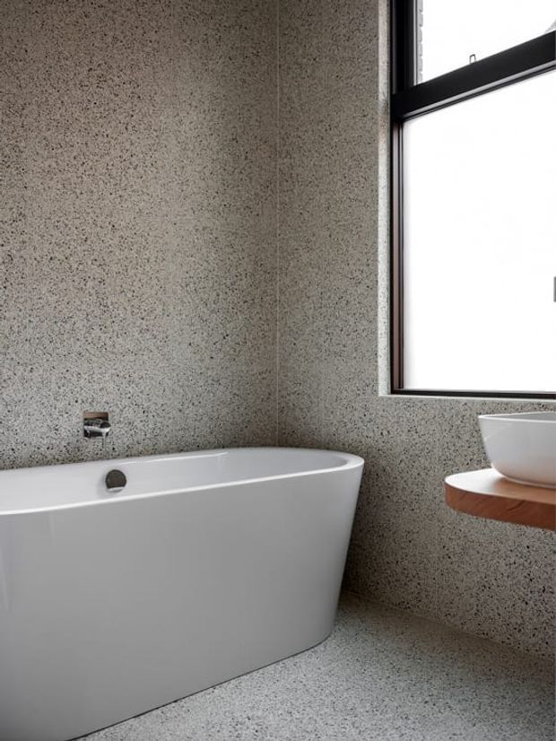 A minimalist bathroom featuring a freestanding white bathtub, a white vessel basin on a curved timber shelf, and dark-framed window glazing. The walls and floor are clad in a continuous grey speckled terrazzo-like finish, creating a restrained contemporary aesthetic.