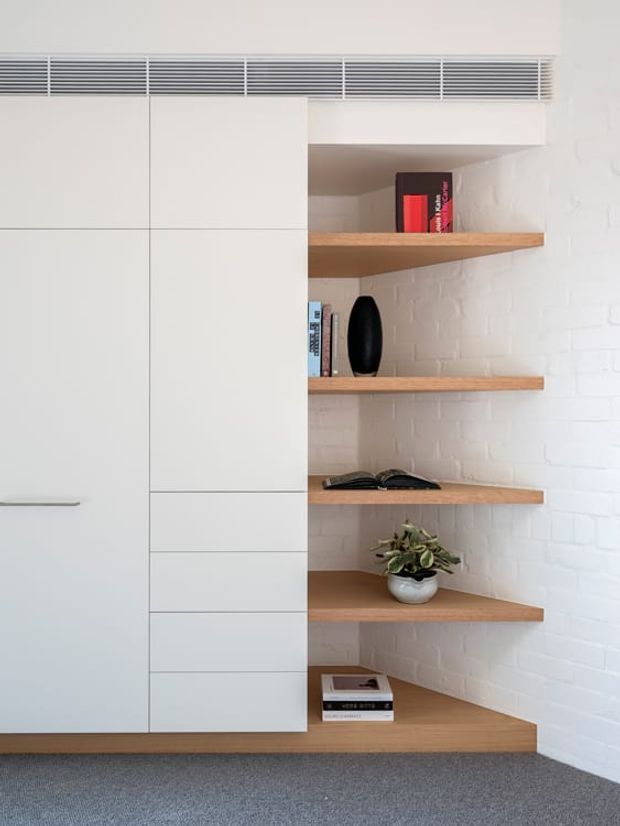 Built-in white cabinetry beside a series of open timber shelves set against a painted white brick wall. The compact joinery includes concealed storage, a long horizontal ventilation grille above, and styled shelving with books and a small plant. Grey carpet covers the floor.