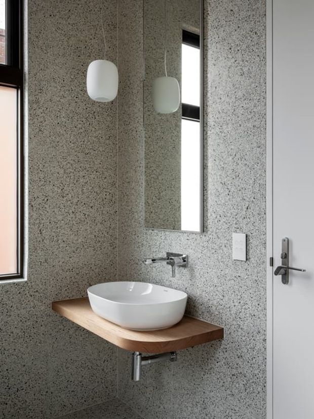 Compact contemporary bathroom with a wall-mounted white vessel basin on a thick natural timber vanity shelf, a wall-mounted chrome tap, speckled grey terrazzo-like wall and floor surfaces, a tall mirrored cabinet or mirror, black-framed window, and a white door with dark hardware. A rounded white pendant light hangs above the vanity.