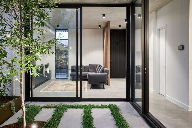 A refined contemporary interior courtyard or entry space with floor-to-ceiling black-framed glazing, pale stone or concrete paving, and a planted garden strip in the foreground. Through the glazing is a minimalist living room with a grey sofa, light flooring, dark vertical timber or ribbed wall panelling, and a warm timber divider. A white hallway with a flush door is visible on the right.