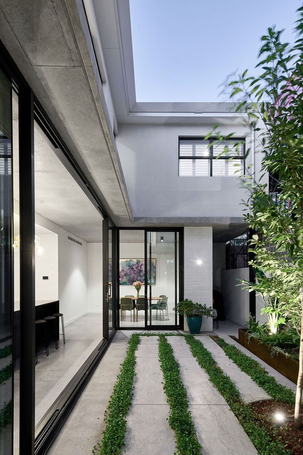 A contemporary internal courtyard viewed from within a home, featuring concrete paving strips with narrow planted bands, lush greenery, black-framed glazed sliding doors, and a high-level open skylit void. The surrounding architecture uses smooth light-grey walls, exposed concrete elements, and large areas of black-framed glazing, creating a bright urban courtyard connection.