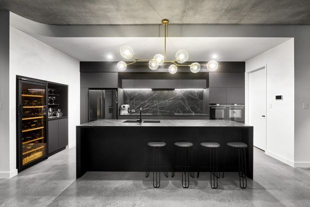 Contemporary monochrome kitchen with a large black island and four stools, dark integrated cabinetry, marble-look splashback, concealed appliances, wine fridge, and sculptural brass-and-glass pendant lighting. Concrete-look flooring and ceiling create an industrial character, balanced by white walls and a bright, open layout.