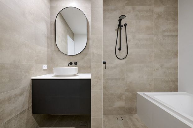 Contemporary bathroom featuring large-format beige-grey stone-look wall and floor tiles, a matte black floating vanity with white vessel basin, rounded black-framed mirror, wall-mounted black tapware, and a walk-in shower beside a white bathtub.