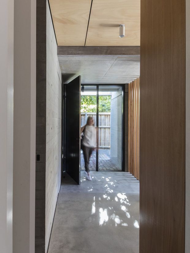 A narrow, minimalist interior passage or entry hall leading to a glazed exterior door. The space features a polished grey concrete floor and ceiling, pale timber ceiling panels, dark exposed structural beams, a textured light masonry wall, and a full-height timber slatted partition or cabinetry along the right side. Natural light and foliage enter through the glazed doorway, with a blurred person moving through the hall.