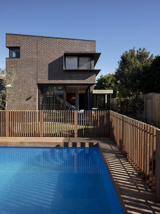Exterior view of a contemporary multi-level brick house overlooking a rectangular swimming pool. The building has a restrained dark brick facade, black-framed glazing, projecting box-like window hoods, and a flat or very low-pitched roofline. Timber slat fencing and decking surround the pool, with strong afternoon shadows across the paved poolside area.