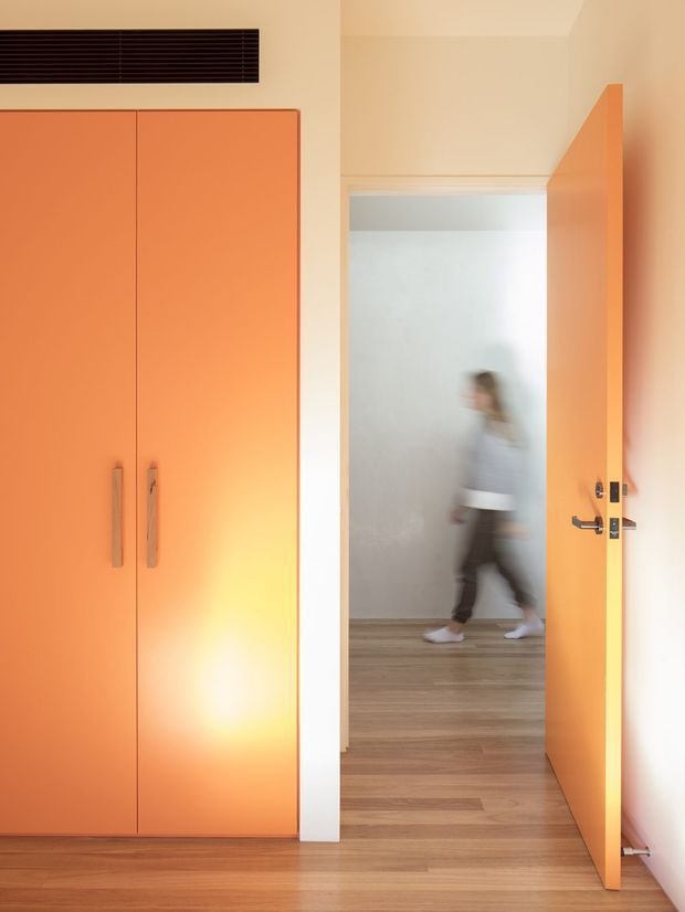 Interior view of a warm, minimalist transitional space with a vivid orange built-in floor-to-ceiling cupboard on the left and a matching orange open door on the right. The doorway opens to a bright hallway with timber flooring; a blurred person is passing through the background. Dark horizontal ventilation or louvre detailing is mounted above the cabinetry, and black door hardware provides contrast.