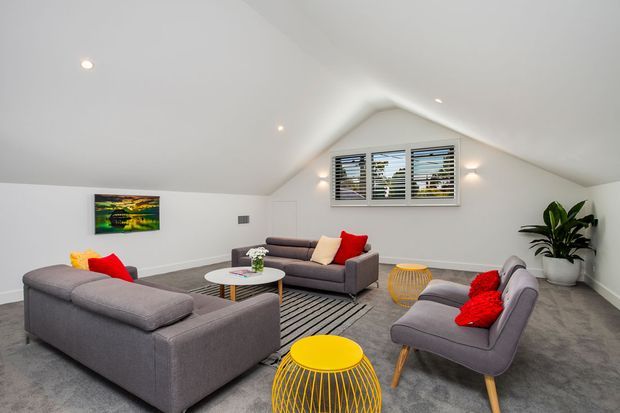 Bright attic-style living room with a steep sloped ceiling, white walls, grey carpet, and a central louvre window. The room is furnished with a large grey sectional sofa, matching armchairs, colourful red and yellow cushions, small round tables, recessed spotlights, and a potted plant.