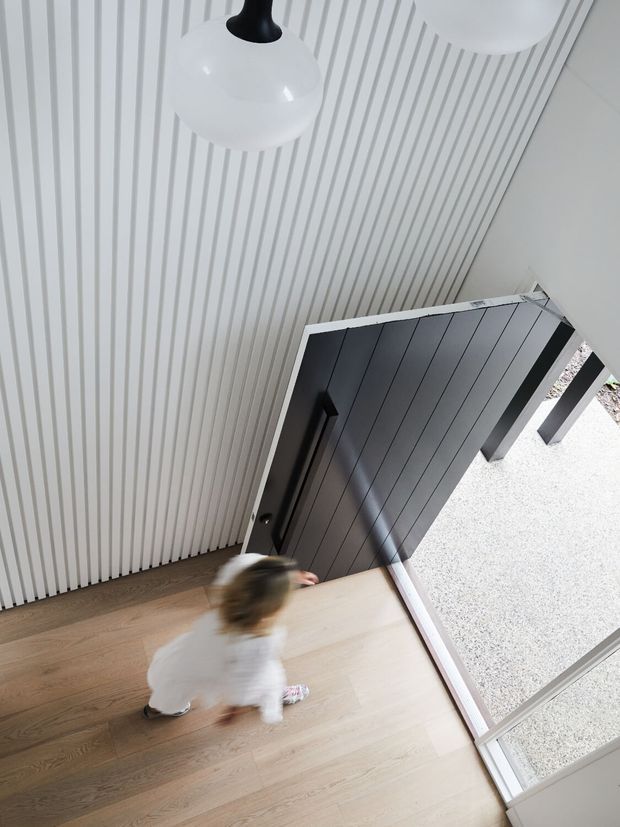 A high-angle view of a bright residential entryway with pale timber flooring, a white vertically ribbed wall, a dark charcoal front door, and large glazed sidelights or entry glazing. Two white pendant lights hang overhead, and a blurred child is moving across the floor.