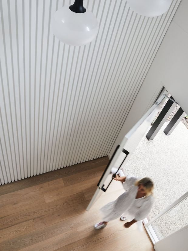A bright contemporary entryway viewed from above, featuring warm timber flooring, a full-height white vertical slatted wall, a large glazed doorway, and sculptural white pendant lights. A person is entering through the door.