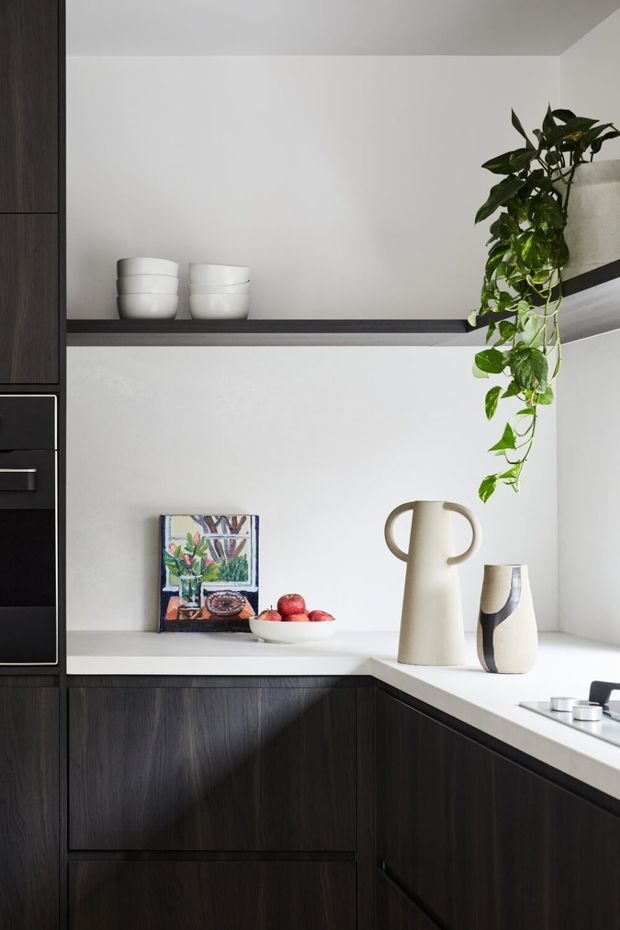 A refined contemporary kitchen corner with dark timber-grain cabinetry, white benchtops and backsplash, open shelving, and integrated appliances. Minimal ceramic vessels, artwork, fruit, and a trailing indoor plant add warmth and texture.