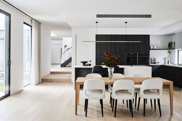Bright contemporary open-plan dining and kitchen area with a light timber dining table, four upholstered dining chairs, pale timber flooring, and a dark built-in kitchen wall with integrated appliances. Full-height glazing and sheer curtains bring in abundant natural light, while a short stairway and hallway are visible beyond.