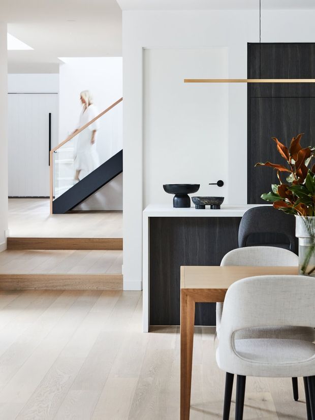Bright contemporary interior showing an open-plan dining and entry area with pale timber flooring, a dark built-in sideboard, minimalist decorative objects, a light timber dining table, upholstered dining chairs, and a black-and-timber staircase visible beyond.