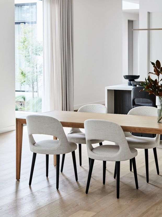 Bright contemporary dining area with a large light timber dining table, sculptural upholstered dining chairs, pale timber flooring, white walls, full-height curtains and generous glazing. A minimalist console and dark accents sit behind the dining setting, with indoor greenery adding an organic touch.