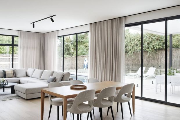 Bright open-plan living and dining area with floor-to-ceiling black-framed glazing, sheer full-height curtains, a large light sectional sofa, and a timber dining table surrounded by upholstered chairs. The space opens onto a pale outdoor patio and landscaped courtyard.
