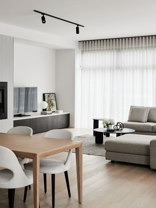 Bright contemporary open-plan living and dining area with a neutral palette. The room features light timber flooring, a timber dining table with upholstered chairs, a dark built-in media cabinet, sheer full-height curtains, a grey sectional sofa, black round coffee tables, and minimalist black track lighting. A television and framed artwork are integrated into the media joinery.