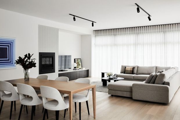 Bright open-plan contemporary living and dining area with a large timber dining table and upholstered chairs in the foreground, a grey sectional sofa and low coffee table to the right, and a minimalist linear fireplace built into a pale feature wall. Full-width sheer curtains, track lighting, pale timber flooring, and restrained neutral furnishings create a clean, airy interior.