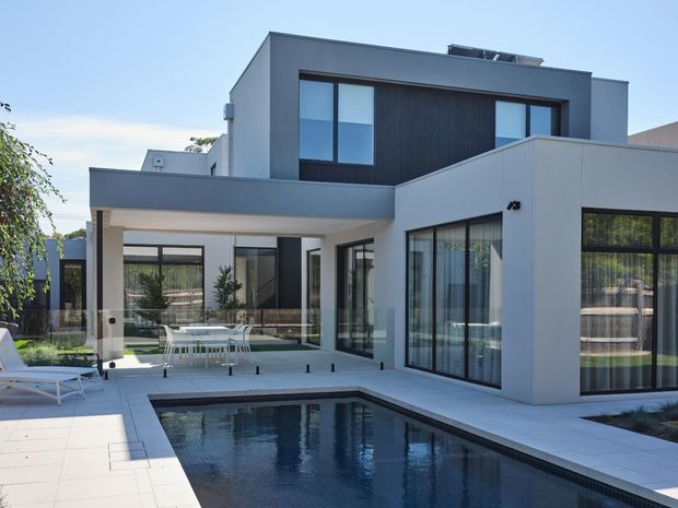 Contemporary multi-level white-rendered residence with dark vertical feature cladding, extensive floor-to-ceiling glazing, and a covered outdoor entertaining area overlooking a rectangular swimming pool. The scene includes a tiled pool terrace, glass balustrade, outdoor dining furniture, and lounge chairs in a bright landscaped setting.