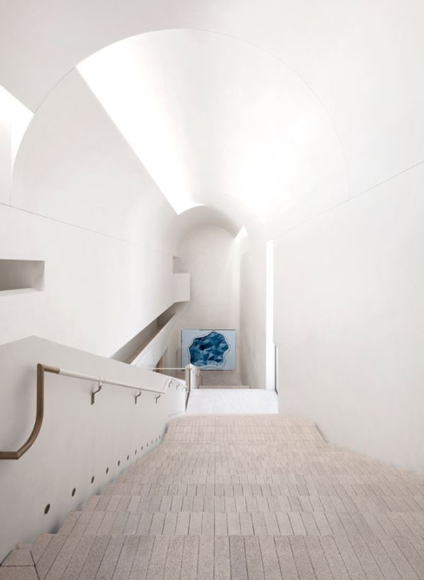 A bright, minimalist stairway descending through a sculptural white interior. The space features a broad curved vaulted ceiling, smooth white walls, pale textured stair treads, and a simple timber-toned handrail with metal supports. A small blue artwork is visible at the lower landing, adding a restrained colour accent.