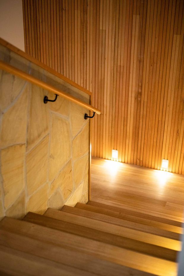 A warmly lit interior stairway descends beside a pale natural-stone wall with a timber handrail and dark metal brackets. The stair treads and landing are timber, while the tall wall is clad in narrow vertical timber slats. Small recessed floor-level lights illuminate the landing and create a calm, sauna-like atmosphere.