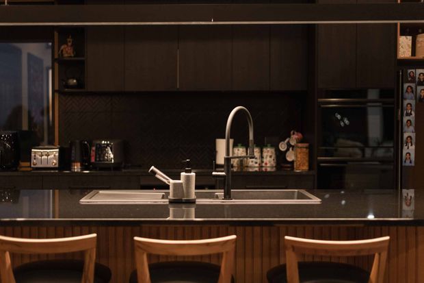 A dark, sophisticated contemporary kitchen viewed across a central island with three timber bar stools. The kitchen features extensive dark cabinetry, a black textured chevron backsplash, a glossy dark stone island benchtop, an undermount sink with a tall gooseneck tap, integrated appliances, and warm timber vertical panelling on the island base. Warm, low lighting creates an intimate and refined atmosphere.