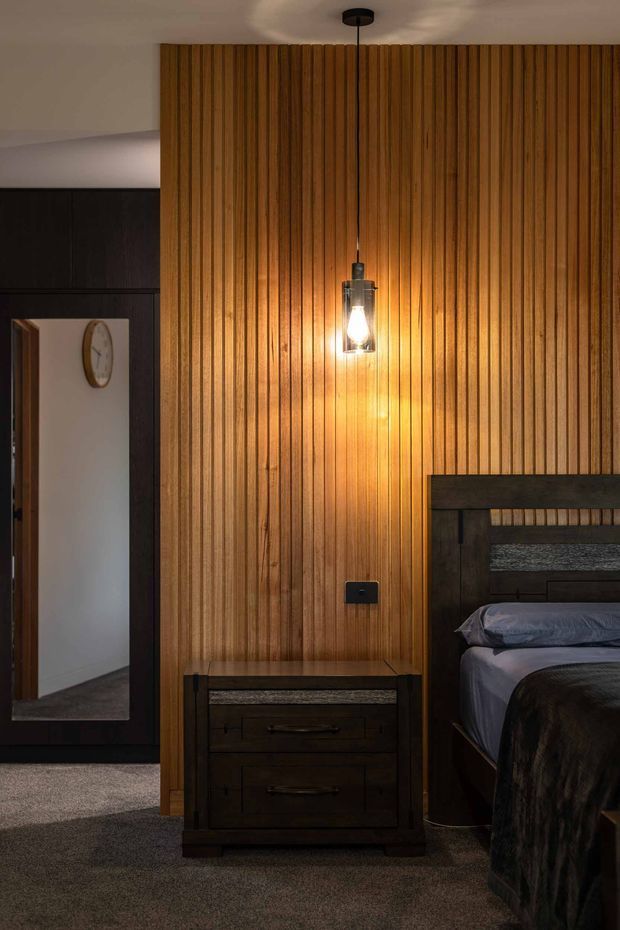 A warmly lit bedroom featuring a dramatic vertical timber-slat feature wall, dark built-in cabinetry with a full-height mirror, and a dark timber bedside table beside a partially visible bed. A pendant light creates a soft amber glow against the timber wall, while carpeted flooring and deep neutral furnishings give the room a moody, refined character.