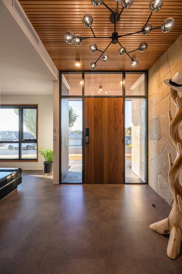 A contemporary residential entry foyer viewed from inside, featuring a tall timber pivot front door with sidelights and a glazed transom. The foyer has a dark polished floor, exposed natural stone feature wall, warm slatted timber ceiling, black-framed glazing, and a sculptural branch-like timber element. A dramatic multi-arm globe pendant light hangs centrally, while a window and partial living area are visible to the left.