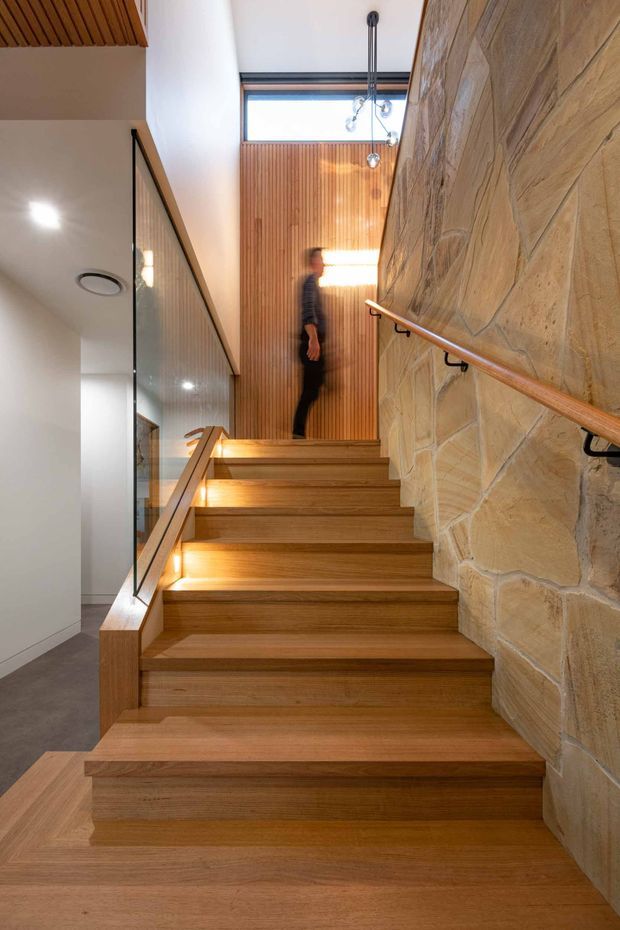 Interior view looking up a timber staircase. The stairs and handrails are warm wood, with integrated step lighting. A rough-faced natural stone wall runs along the right side, while the opposite side has a clear glass balustrade. Vertical timber panelling and a high clerestory window illuminate the upper landing. The space has a refined contemporary character with strong natural materials and architectural detailing.