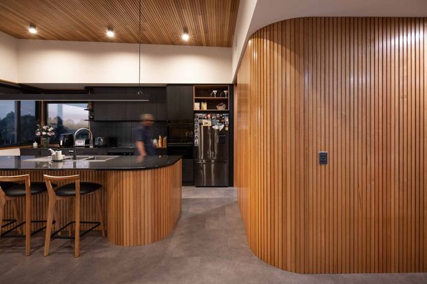 A contemporary open-plan kitchen dominated by warm vertical timber battens. The space features a curved timber-clad island with dark benchtops, integrated sink and seating, matte-black cabinetry and splashback, stainless-steel appliances, and a matching curved timber feature wall. A timber slatted ceiling, recessed lighting, and polished concrete-look flooring complete the refined, warm interior.