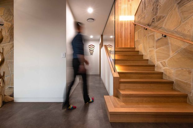 A contemporary interior hallway and stairway viewed from the lower level. Broad timber stairs rise alongside a floor-to-ceiling glass balustrade, with warm vertical timber slats and a dramatic natural stone feature wall. The adjoining corridor has smooth pale walls, recessed ceiling lights, and a dark polished floor. A blurred person provides a sense of scale.