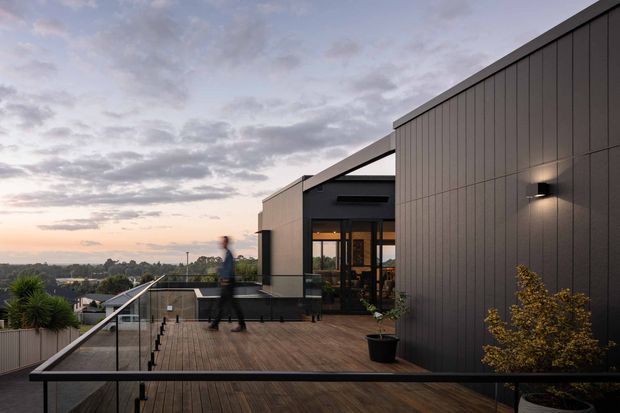 A contemporary rooftop terrace or elevated deck beside a dark-clad building, featuring timber decking, frameless glass balustrades with dark handrails, architectural wall lighting, potted plants, and glazed doors leading into the building. The elevated view overlooks a suburban landscape beneath a cloudy sunset sky.