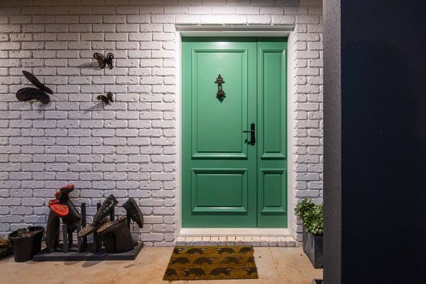 A decorative entryway featuring a vivid green double-panel front door with traditional raised moulding, black hardware, and a small ornamental door knocker. The door is set within a white-painted exposed brick wall, with insect-themed wall decorations, potted plants, footwear, and a patterned doormat on a pale hard-surface floor. The scene is warmly lit and visually eclectic.