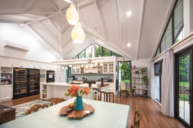 Bright open-plan kitchen and dining space with a high vaulted white ceiling, exposed painted timber trusses, clerestory glazing, and large black-framed windows and doors. The room features white built-in kitchen cabinetry with glass-front display storage, a large island, dark wood flooring, a pale dining table, pendant lighting, and a relaxed traditional country-inspired fit-out. Lush greenery is visible outside through the glazing.