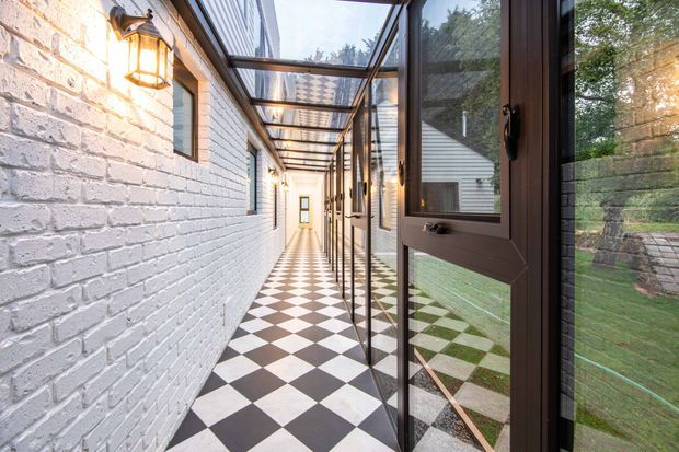A narrow enclosed passageway with a striking black-and-white checkerboard tile floor, white-painted exposed brick walls, and a glazed pitched canopy overhead. Dark timber-framed glass doors and windows line the right side, reflecting and opening toward a landscaped garden. Warm wall lanterns illuminate the corridor, creating a refined traditional-contemporary architectural transition space.
