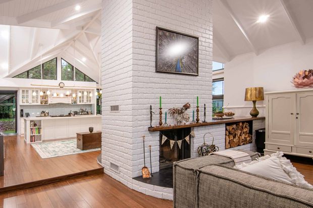 Bright open-plan living room with a prominent white-painted brick fireplace chimney breast, traditional mantel and hearth, exposed white timber roof structure, and warm timber flooring. A textured sectional sofa faces the fireplace, while built-in shelving and cabinetry are visible in the adjoining area. Large triangular clerestory-style glazing brings in natural light, creating an airy yet warm interior.
