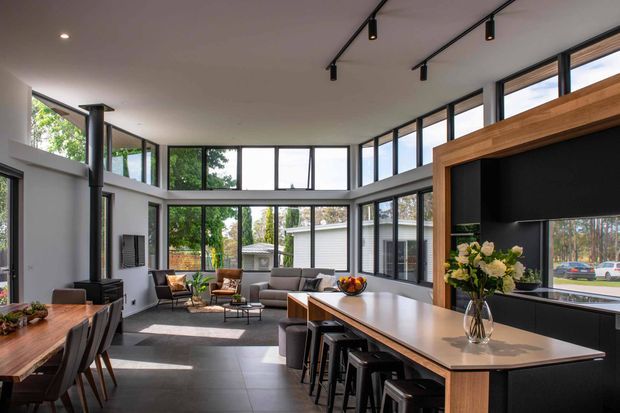 Bright, spacious open-plan contemporary interior combining a kitchen, dining area and living room. The room features extensive black-framed windows, a high ceiling with track lighting, dark tiled flooring, a large island with seating, timber-framed black cabinetry, and a relaxed lounge area around a fireplace or media feature. Abundant glazing brings in greenery and daylight.