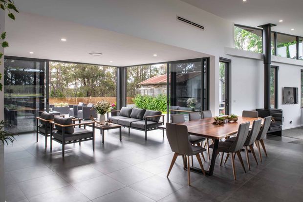 A bright, open-plan contemporary living and dining area with floor-to-ceiling glazing, dark tiled flooring, white walls, and a high ceiling. A dining table with upholstered chairs occupies the foreground, while a lounge seating arrangement sits beside expansive windows. A black freestanding wood-burning stove and flue provide a strong feature, with greenery and an outdoor setting visible beyond the glazing.