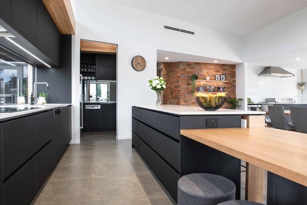 Contemporary open-plan kitchen with a large matte-black island and cabinetry, integrated timber breakfast bar, concrete-look flooring, white walls, and a contrasting exposed brick feature wall with open shelving. A dining area and secondary kitchen zone are visible beyond the island.