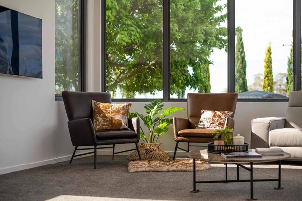 A bright, contemporary living room with floor-to-ceiling dark-framed windows overlooking leafy greenery. The room features two brown leather lounge chairs, a neutral upholstered sofa, a black metal and timber coffee table, a potted plant, patterned cushions, a textured rug, and grey carpet. A blue abstract artwork hangs on the pale wall.