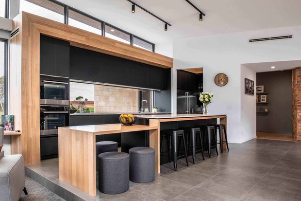A bright, contemporary open-plan kitchen with a large timber-framed black cabinetry wall, integrated double ovens, dark stone-look benchtops, and a long island with bar seating. The room has high clerestory-style glazing, track lighting, a tiled floor, and an opening toward a darker hallway or living area with exposed brick detailing.