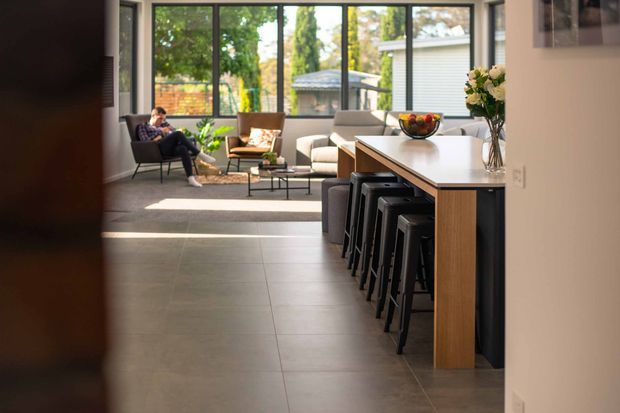 A bright, open-plan contemporary interior viewed from a tiled circulation area toward a living space and kitchen island. The kitchen features a long light benchtop with timber cabinetry and dark bar stools, while the living area has lounge seating, a low coffee table and large black-framed windows overlooking greenery. Natural light, neutral finishes and warm timber accents create an inviting atmosphere.