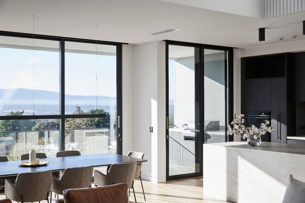 Bright contemporary open-plan kitchen and dining area with expansive black-framed glazing overlooking a distant landscape. A large dining table with upholstered chairs sits beside the windows, while a dark built-in kitchen with a substantial pale stone island occupies the right side. The space has light timber flooring, white walls, strong natural light, and a clear indoor-outdoor connection through the glazed door.