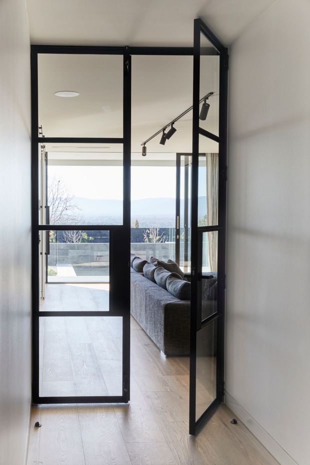 A narrow interior passage with pale timber flooring and white walls, looking through a black-framed glazed steel-style door into a bright living room. The open door and matching glazed partition create a strong industrial-modern feature, with a grey sofa, track lighting, expansive glazing, and an outdoor terrace visible beyond.